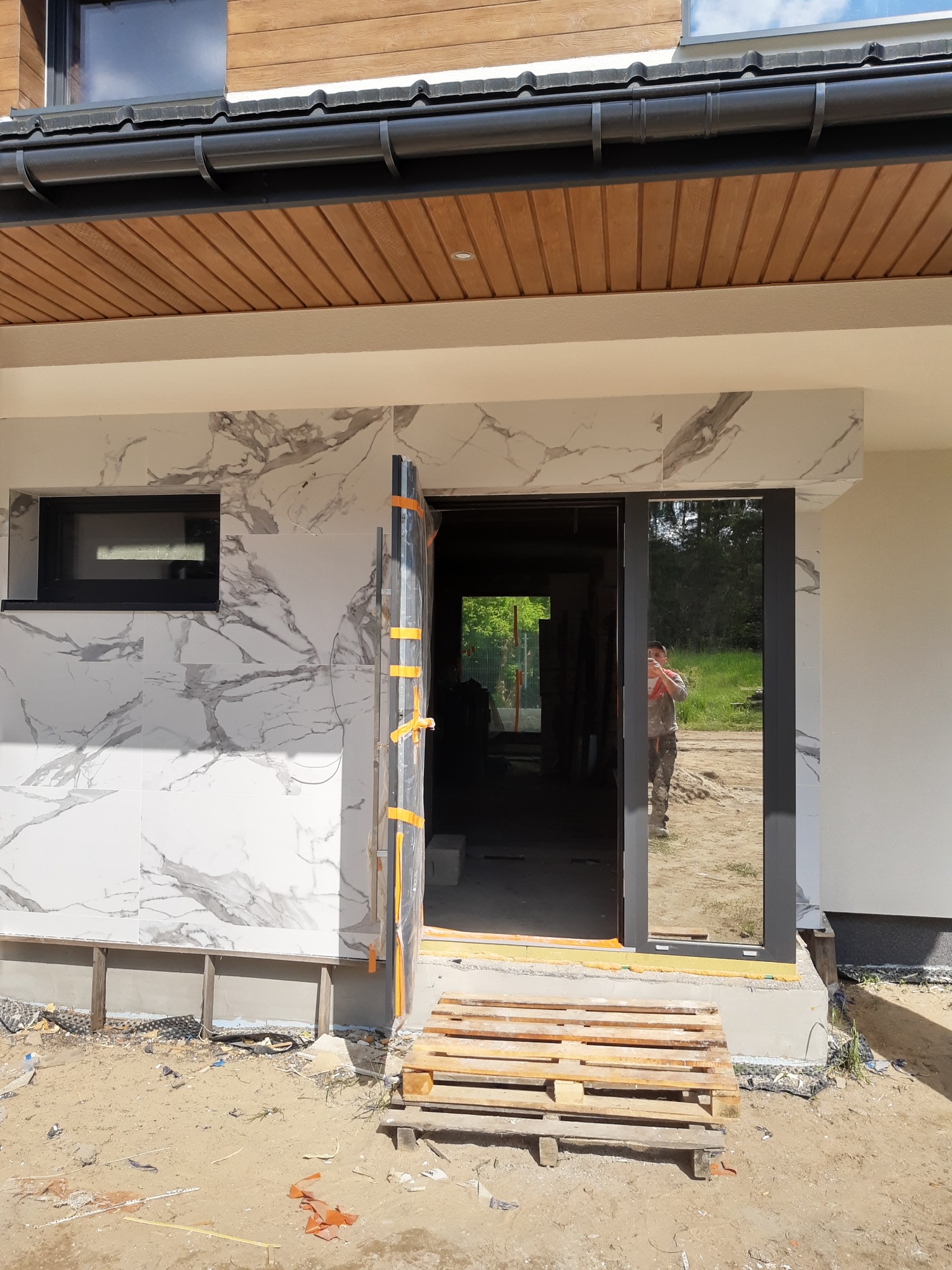 Montaż elewacji z płytek imitujących marmur, widoczne okno z czarną ramą i drzwi z folią ochronną, paleta drewniana na ziemi, odbicie osoby w szybie.