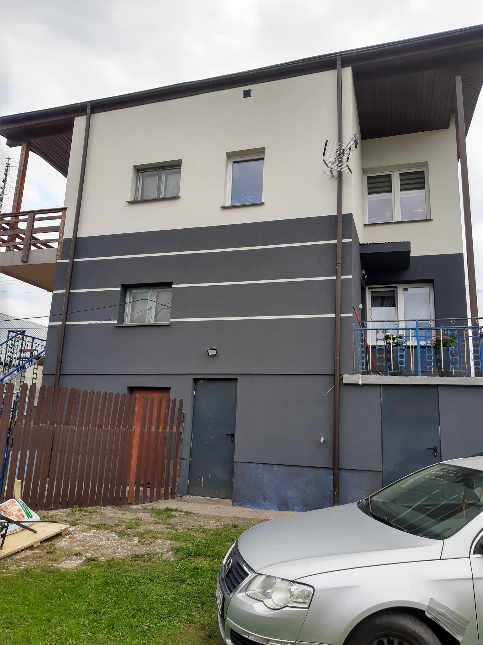 Dwukondygnacyjny dom z elewacją w odcieniach szarości i bieli, z balkonem z niebieską balustradą i anteną satelitarną, widok z poziomu trawnika.