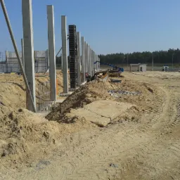 Budowa hali: betonowe słupy konstrukcyjne w trakcie montażu, widoczne szalunki i zbrojenia, w tle sprzęt budowlany i teren przygotowany pod wylewkę.