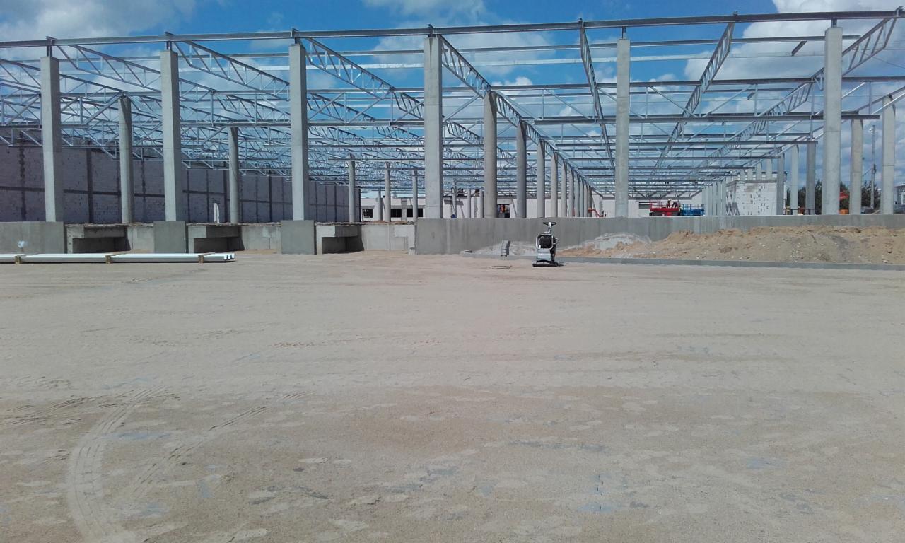 Wczesny etap budowy hali z widoczną stalową konstrukcją szkieletową, betonowymi słupami, przygotowanym podłożem i zagęszczarką wibracyjną na placu budowy.
