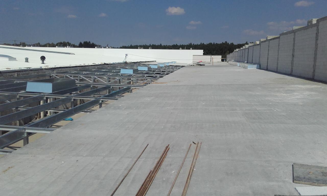 Wnętrze hali w trakcie budowy, widoczne stalowe konstrukcje nośne i fragmenty betonowej posadzki w surowym stanie, w tle ściany z betonu komórkowego i białe elementy dachu.