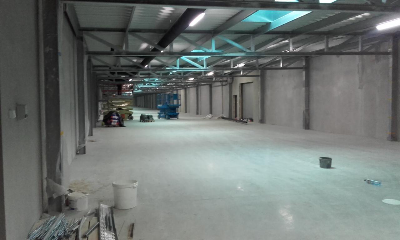 Wnętrze hali w trakcie budowy, widoczne surowe ściany, stalowa konstrukcja dachu, podnośnik koszowy, wiaderka z farbą i drobne narzędzia na posadzce.