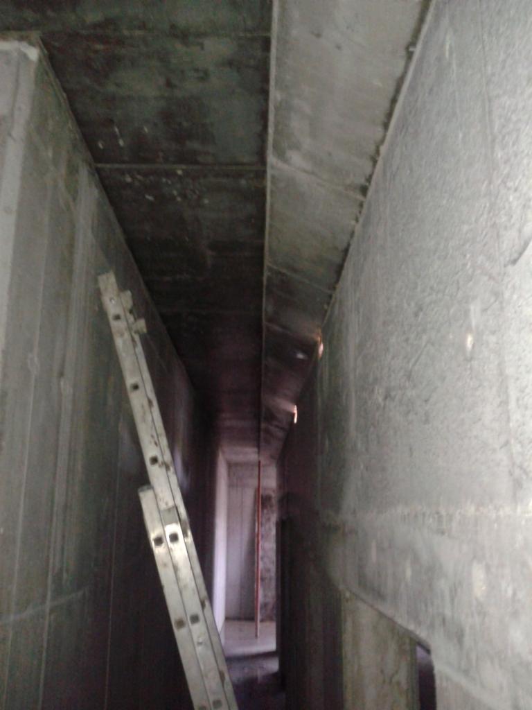 Wąskie, surowe wnętrze betonowej konstrukcji w trakcie budowy, z drabiną opartą o ścianę i widocznym prześwitem na końcu korytarza, oświetlone słabym światłem.