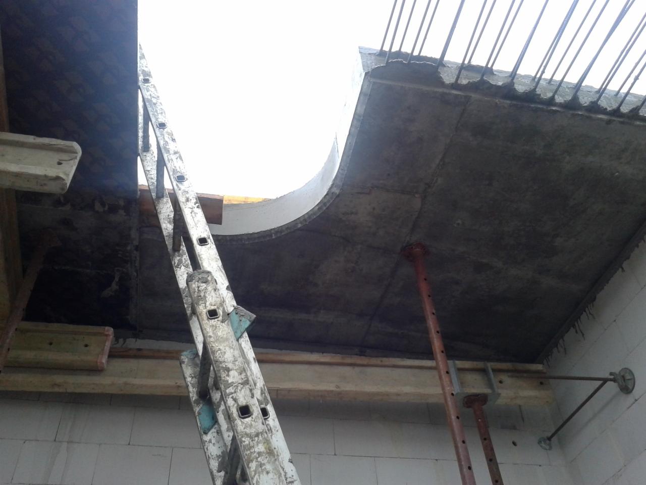 Wnętrze w trakcie budowy z widocznym betonowym stropem podpartym stemplami, szalunek z otworem w kształcie łuku i drabina oparta o ścianę z białych bloczków.