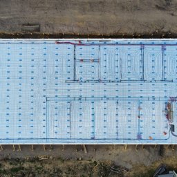 PRECYZJA - Widok z góry na przygotowaną płytę fundamentową z izolacją i rozłożonym systemem ogrzewania podłogowego, z widocznymi rurami i elementami montażowymi, otoczoną ziemią i materiałami budowlanymi.
