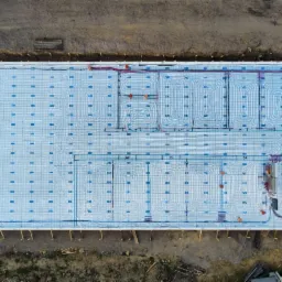 Widok z góry na przygotowaną płytę fundamentową z izolacją i rozłożonym systemem ogrzewania podłogowego, z widocznymi rurami i elementami montażowymi, otoczoną ziemią i materiałami budowlanymi.