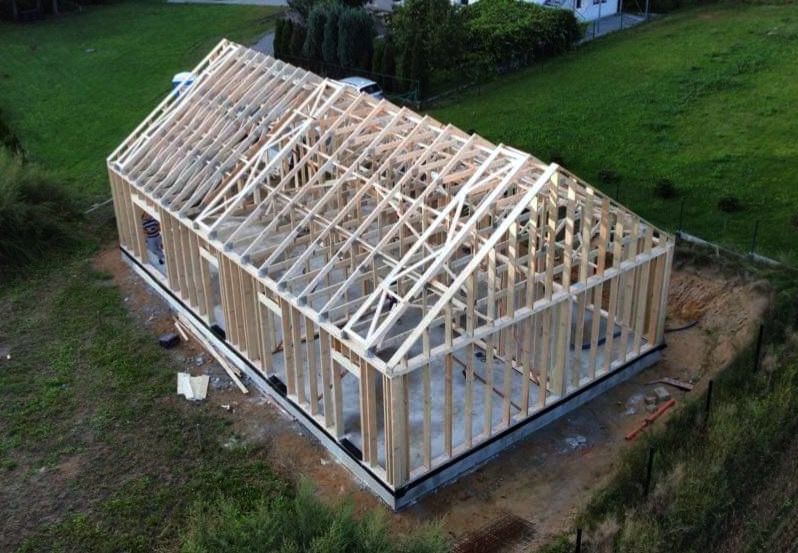 Wznoszenie szkieletu domu z drewnianą konstrukcją dachu, widok z góry na otoczenie z zielenią i fragmentem zaparkowanego samochodu.