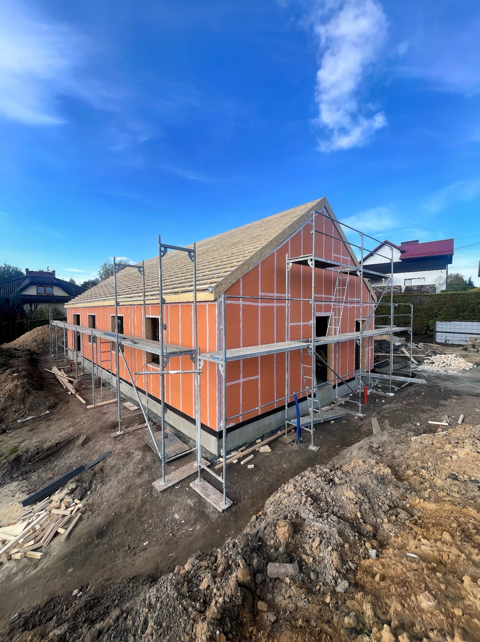 Surowy stan domu szkieletowego z pomarańczową elewacją i drewnianym dachem, otoczony rusztowaniami na tle błękitnego nieba.