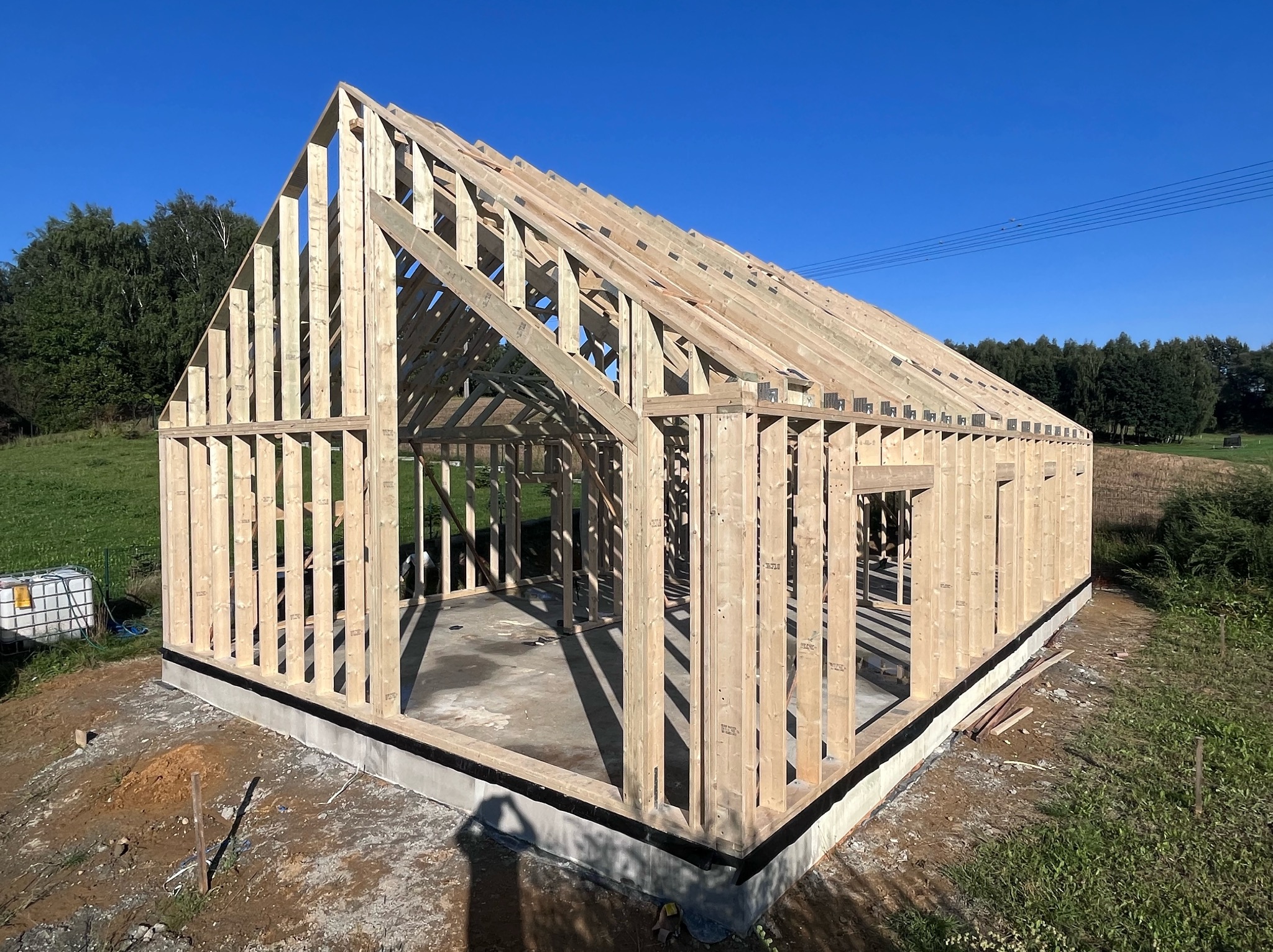 Surowy szkielet domu w trakcie budowy, widoczne drewniane belki i słupy na betonowym fundamencie w słoneczny dzień.