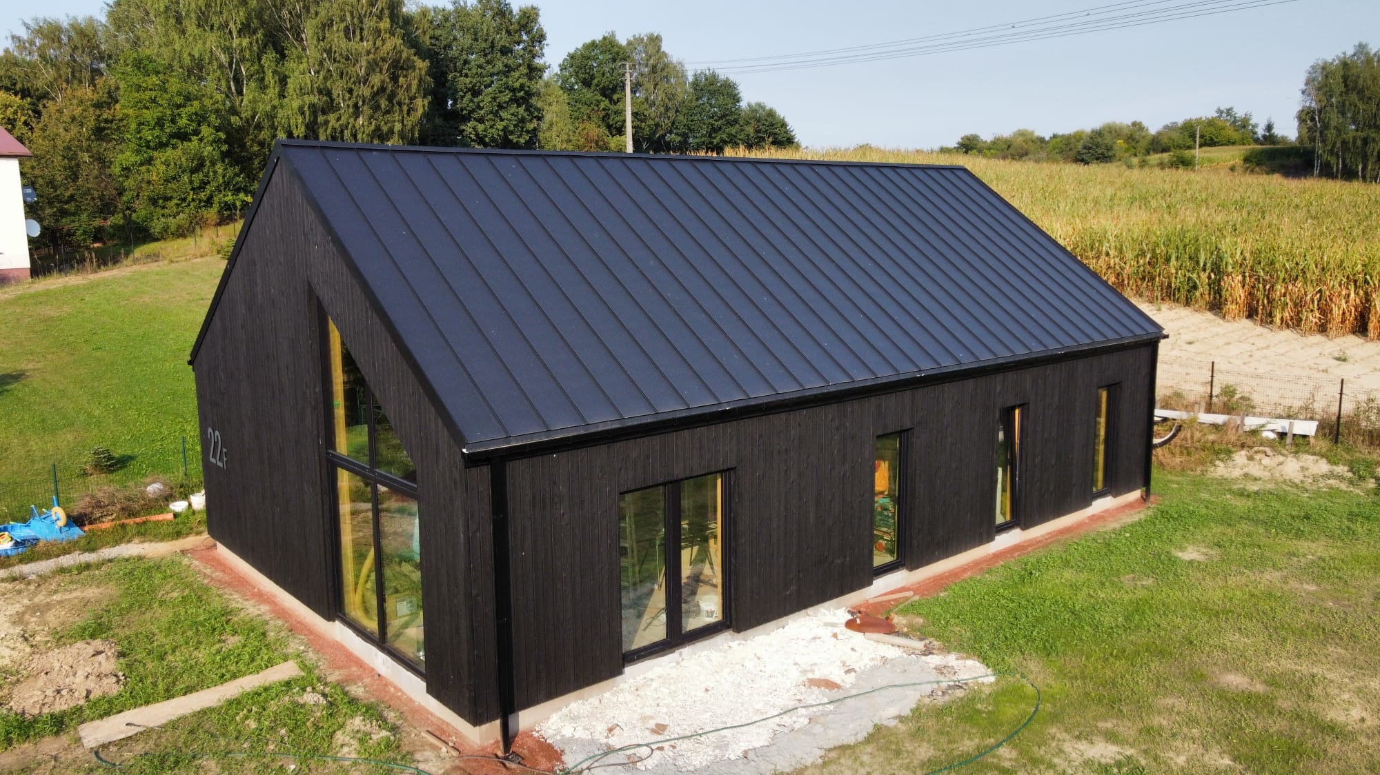 Czarny dom szkieletowy z ciemnym dachem blachodachówką i dużym trójkątnym oknem, otoczony trawnikiem i polem kukurydzy w tle.