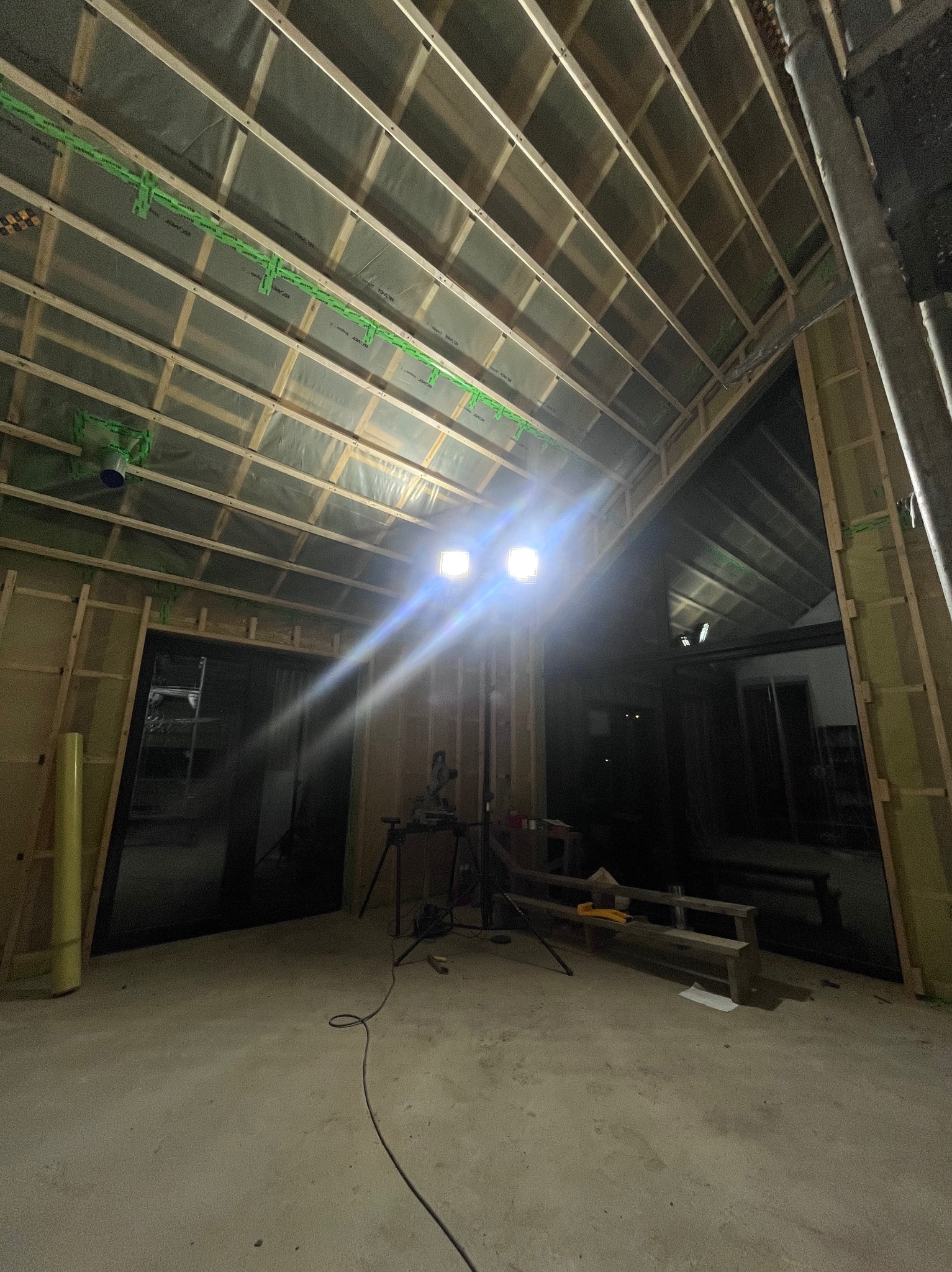 Wnętrze domu szkieletowego w trakcie budowy, widoczne drewniane elementy konstrukcyjne dachu i ścian, ocieplenie, folia paroizolacyjna z zielonymi elementami montażowymi, oświetlenie budowlane...