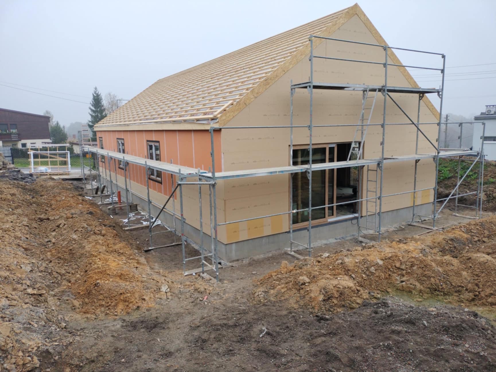Budowa domu szkieletowego: Widok z zewnątrz na częściowo wykończony dom szkieletowy z widocznymi rusztowaniami, odsłoniętą konstrukcją dachu i przygotowanym terenem wokół.