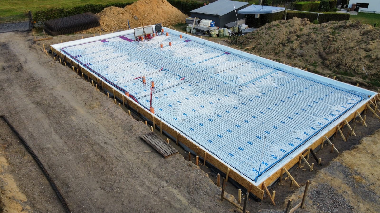 Przygotowana płyta fundamentowa z izolacją i instalacją ogrzewania podłogowego, otoczona drewnianym szalunkiem, widok z góry na plac budowy.