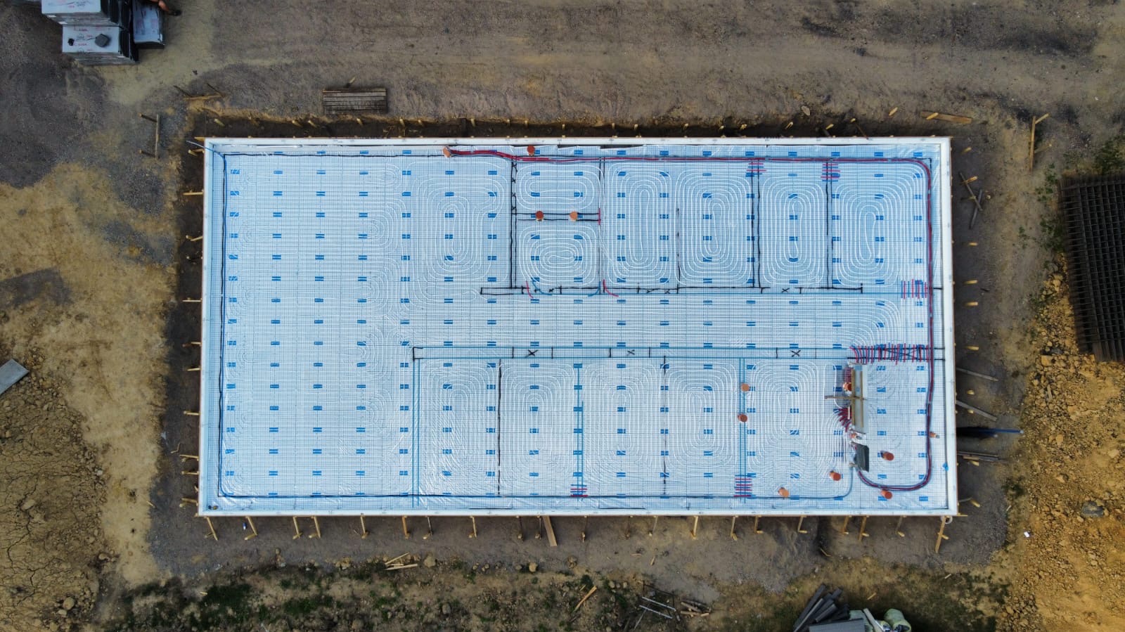 Widok z góry na przygotowaną płytę fundamentową z izolacją i rozłożonym systemem ogrzewania podłogowego, z widocznymi rurami i elementami montażowymi, otoczoną ziemią i materiałami budowlanymi.