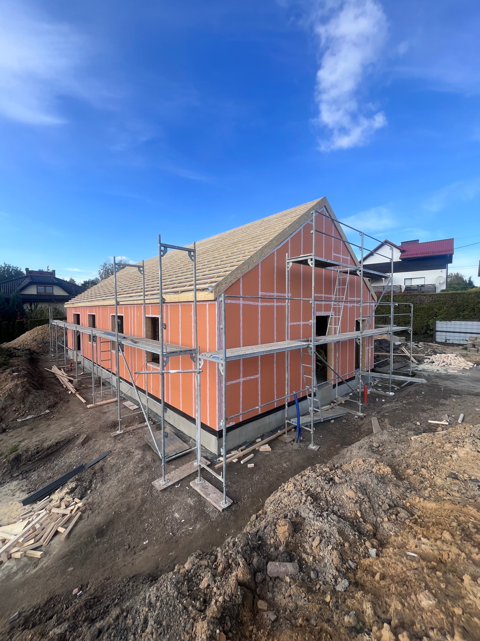 Budowa domu szkieletowego w trakcie: widoczne ściany z izolacją, rusztowania i przygotowany dach z desek na tle błękitnego nieba.