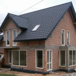 Dwukondygnacyjny dom w trakcie budowy, z widoczną konstrukcją z cegły i zamontowanymi oknami. Czarny dach z dachówkami. Widoczne materiały budowlane na zewnątrz.