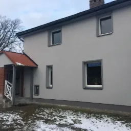 Dwukondygnacyjny dom z szarą elewacją i oranżerią z cegły, widoczny fragment dachu i komina, podłoże częściowo pokryte śniegiem.