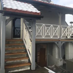 Szare wejście do domu z drewnianymi schodami i białą balustradą, widoczne ślady śniegu, drzwi do piwnicy pod balkonem.