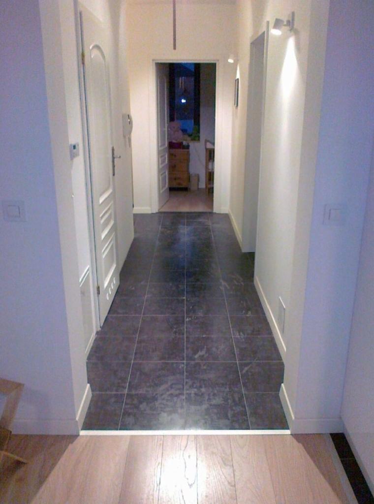 Korytarz z ciemnymi, kwadratowymi płytkami podłogowymi przechodzącymi w jasny drewniany parkiet. Białe ściany z oświetleniem punktowym i widocznymi drzwiami prowadzącymi do innych pomieszczeń.