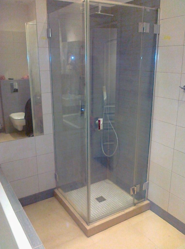 Nowoczesna, narożna kabina prysznicowa z przezroczystego szkła, z widocznym odpływem w kwadratowej kratce na białej, mozaikowej podłodze. W tle widoczne płytki ścienne i fragment toalety.