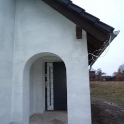 Ryszard Golenia - Wejście do domu w Łubnianach z łukowatym portalem i świeżo otynkowaną elewacją w kolorze złamanej bieli, widoczne przewody pod dachem i fragment zadaszenia nad drzwiami.