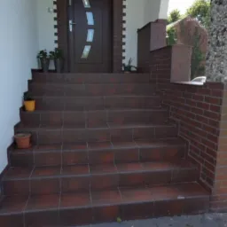 Schody wejściowe wyłożone brązowymi płytkami ceramicznymi, z ceglaną balustradą i kolumną z kamieni ozdobnych, prowadzące do ciemnych drzwi wejściowych.