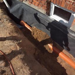 Ryszard Golenia - Wykonanie izolacji pionowej fundamentu z folią kubełkową i ułożoną rurą drenarską w wykopie obok ściany budynku z cegły.