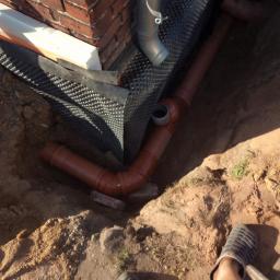 Ryszard Golenia - Odsłonięty fundament budynku z cegły, zabezpieczony folią kubełkową i ocieplony styropianem, widoczne rury kanalizacyjne w wykopie.