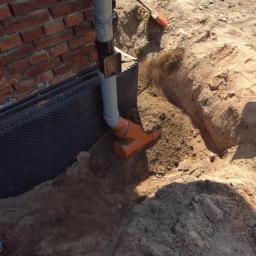 Ryszard Golenia - Izolacja pionowa fundamentu budynku z cegły, zabezpieczona folią kubełkową i rurą odprowadzającą wodę deszczową, widoczne prace ziemne.