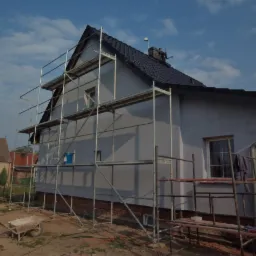 Dom jednorodzinny w trakcie tynkowania elewacji, widoczne rusztowania, fragment odsłoniętego fundamentu z cegły i niebieskie wiaderko na rusztowaniu.