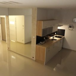 BUKU KRZYSZTOF KOCZYŁO - Widok na nowoczesną kuchnię z drewnianymi szafkami i czarnym blatem, z widoczną częścią salonu i sypialni w tle, oświetloną ozdobną lampą.