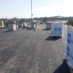 Widok z góry na płaski dach pokryty papą termozgrzewalną, z widocznymi kominami wentylacyjnymi i paletami z materiałem izolacyjnym BMI Icopal, w tle zabudowa miejska i zieleń.