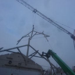 BUKU KRZYSZTOF KOCZYŁO - Demontaż drewnianej konstrukcji dachu za pomocą dźwigu na tle pochmurnego nieba, widoczne elementy starej konstrukcji.