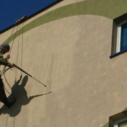 BUKU KRZYSZTOF KOCZYŁO - Pracownik na linach myje elewację budynku z zielonymi, okrągłymi elementami dekoracyjnymi za pomocą myjki ciśnieniowej, widoczny cień pracownika.