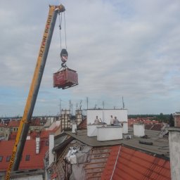 BUKU KRZYSZTOF KOCZYŁO - Żuraw podnosi paletę z dachówkami na dach budynku w trakcie prac dekarskich, widoczni dekarze i częściowo zdemontowana konstrukcja dachu.