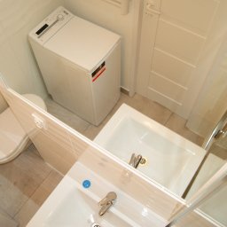 BUKU KRZYSZTOF KOCZYŁO - Umywalka z baterią, toaleta i pralka w małej, wykończonej na biało łazience, widoczne w lustrze.