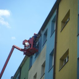 BUKU KRZYSZTOF KOCZYŁO - Dwóch pracowników w czerwonym podnośniku koszowym maluje elewację bloku na wysokości, widoczny fragment elewacji w różnych kolorach: zielonym, żółtym i niebieskim.