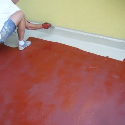 BUKU KRZYSZTOF KOCZYŁO - Mężczyzna w roboczych szortach maluje pędzlem białą listwę przypodłogową na tle żółtej ściany i czerwonej podłogi.