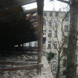 BUKU KRZYSZTOF KOCZYŁO - Demontaż dachu z widocznymi elementami konstrukcyjnymi i pracownikiem w kasku na tle budynku mieszkalnego i drzew