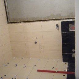 BUKU KRZYSZTOF KOCZYŁO - Wnętrze łazienki w trakcie układania jasnych płytek podłogowych z krzyżykami dystansowymi, widoczna poziomica i czarne płytki na ścianie z przygotowanymi otworami na armaturę.