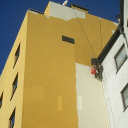 BUKU KRZYSZTOF KOCZYŁO - Pracownik na linach maluje elewację budynku na biało, kontrastując z żółtą, niepomalowaną częścią ściany; widoczny fragment rynny i okna sąsiedniej części budynku, na górze anteny.