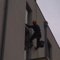 BUKU KRZYSZTOF KOCZYŁO - Pracownik alpinistyczny myje okna na wysokości w nowoczesnym budynku wielorodzinnym, ubrany w kask i uprząż, z wiadrem zawieszonym obok.