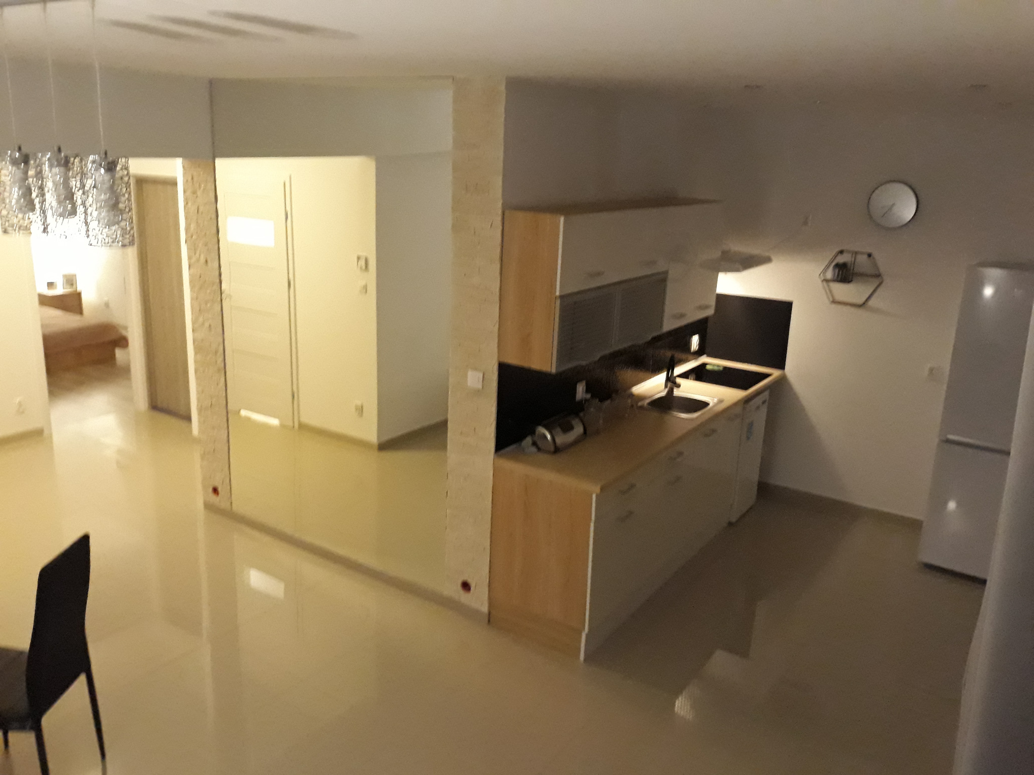 Widok na nowoczesną kuchnię z drewnianymi szafkami i czarnym blatem, z widoczną częścią salonu i sypialni w tle, oświetloną ozdobną lampą.