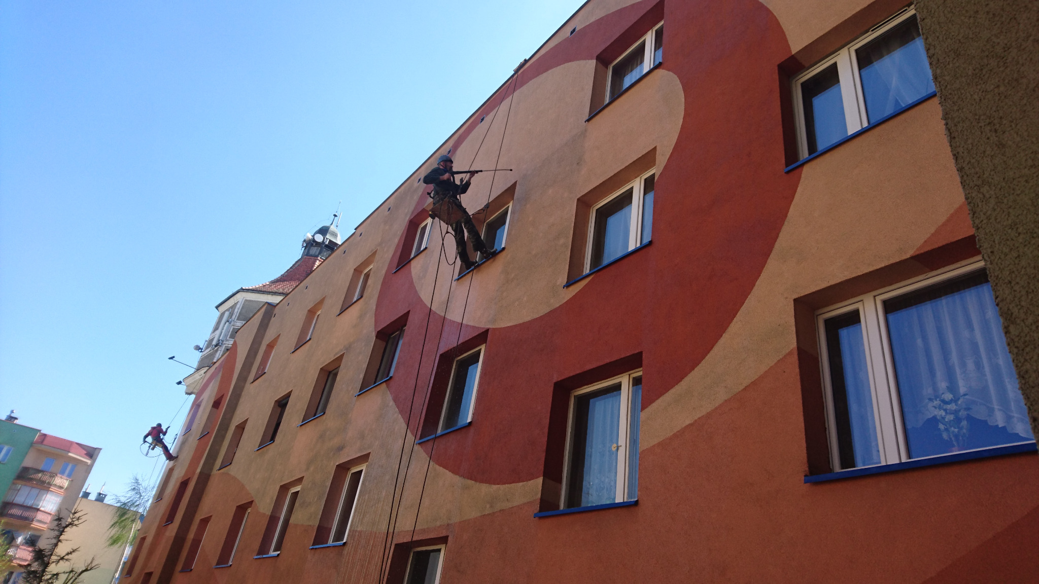 Pracownik na linie alpinistycznej maluje elewację budynku z abstrakcyjnym wzorem, w tle błękitne niebo i sąsiednie budynki.