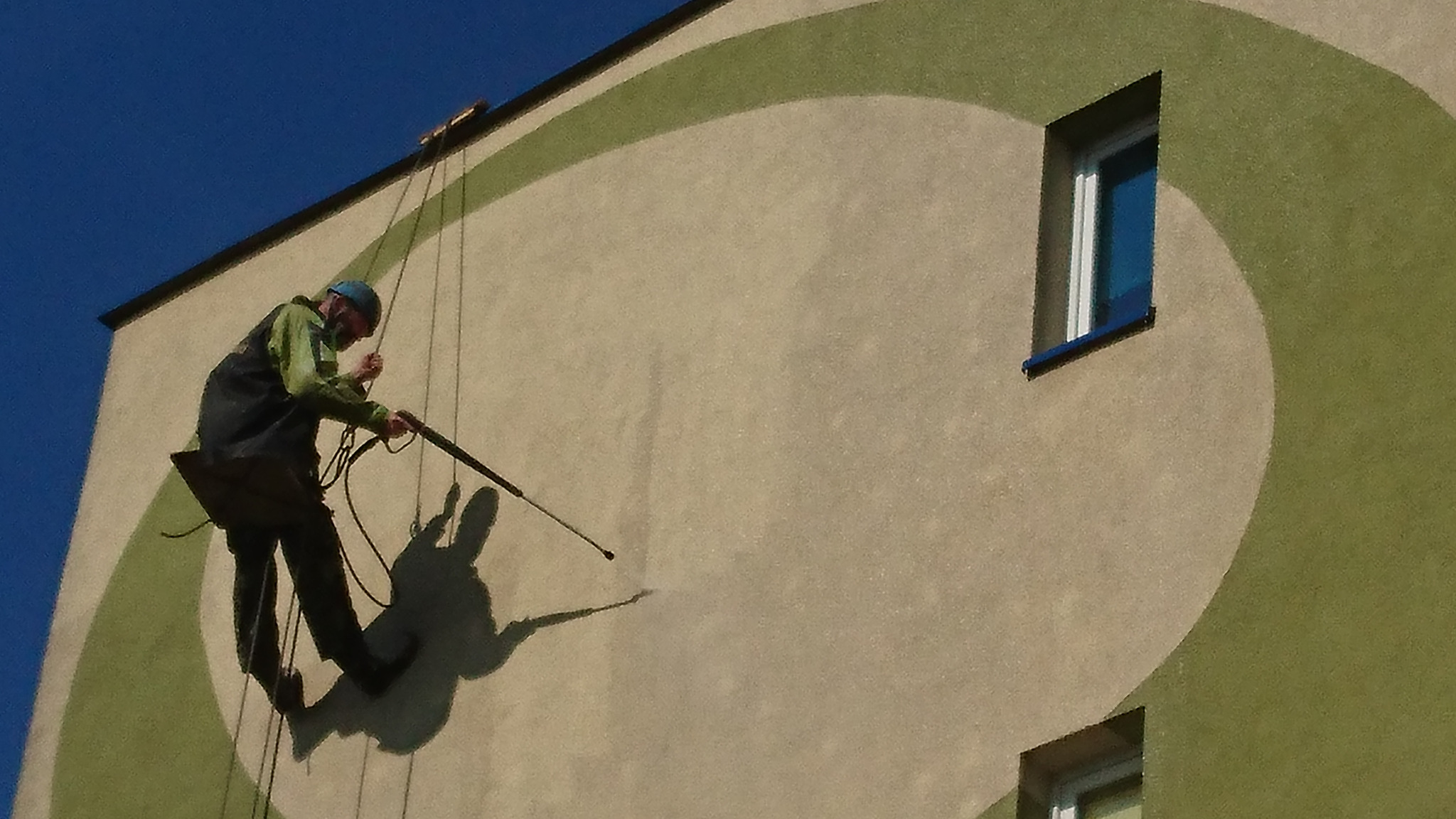 Pracownik na linach myje elewację budynku z zielonymi, okrągłymi elementami dekoracyjnymi za pomocą myjki ciśnieniowej, widoczny cień pracownika.