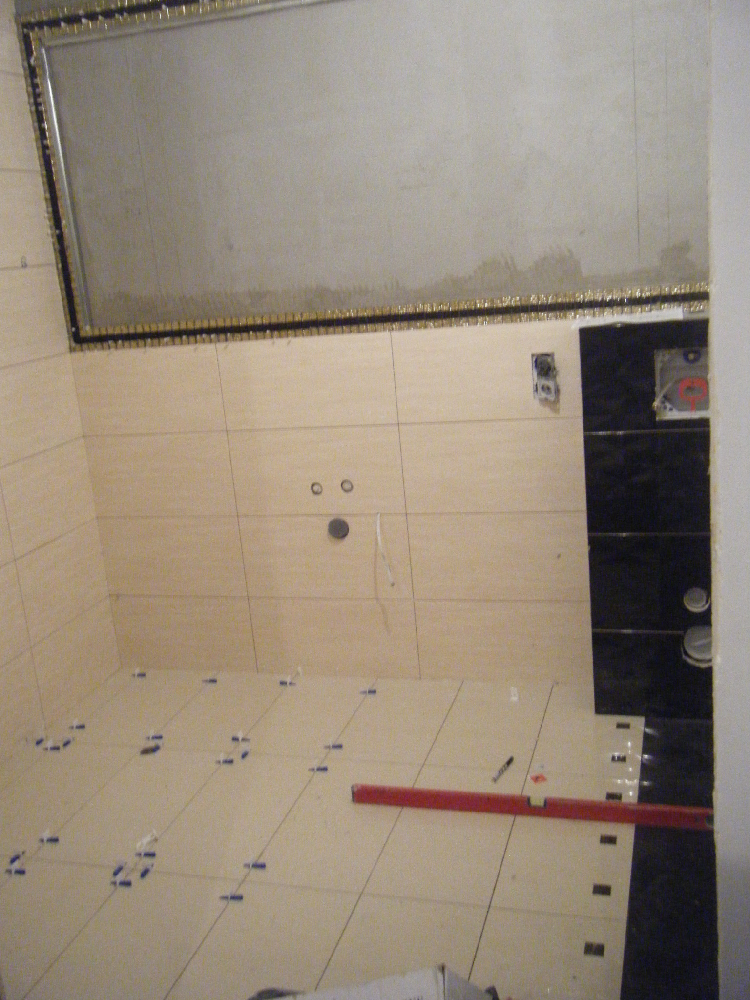 Wnętrze łazienki w trakcie układania jasnych płytek podłogowych z krzyżykami dystansowymi, widoczna poziomica i czarne płytki na ścianie z przygotowanymi otworami na armaturę.