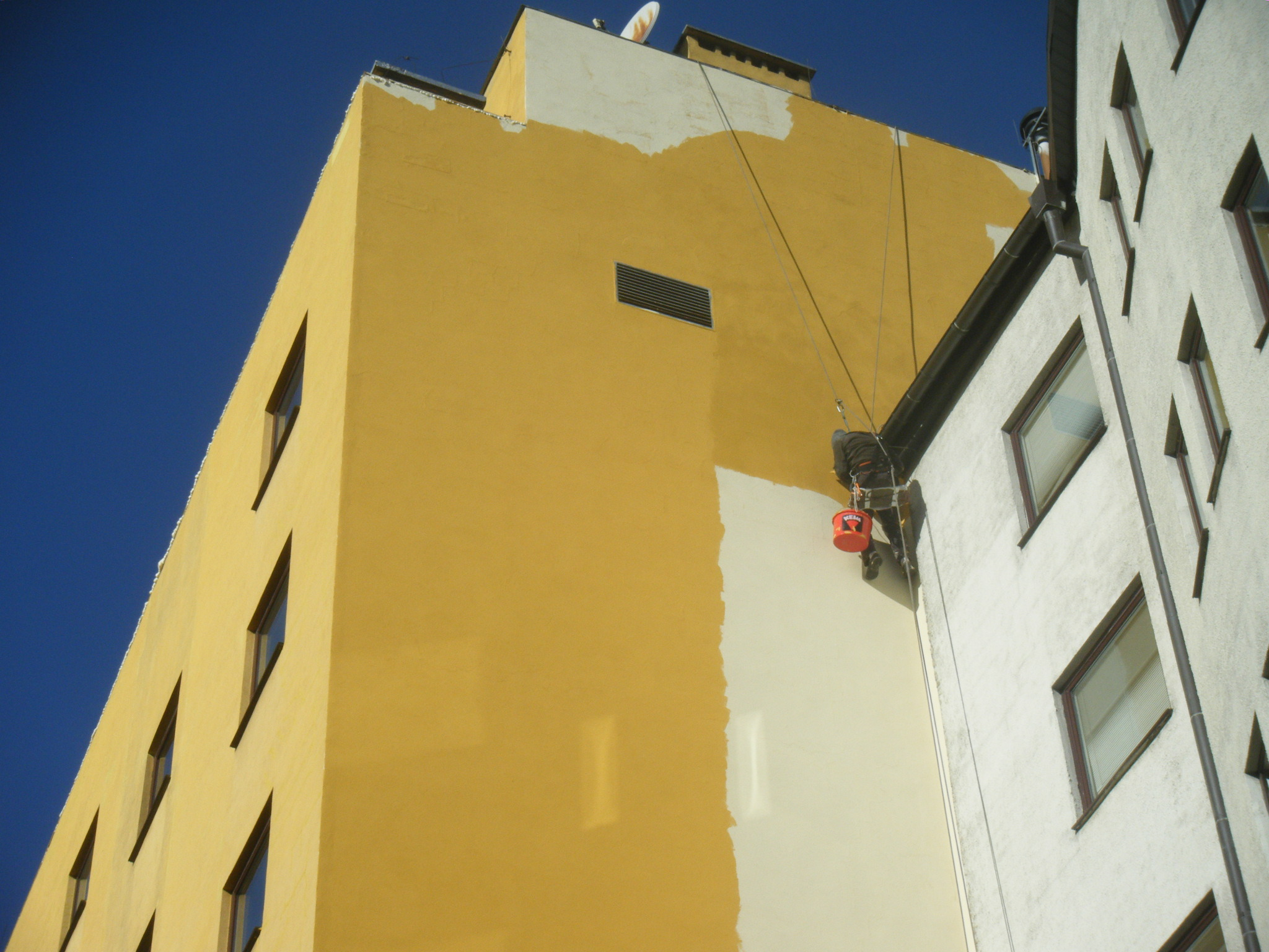 Pracownik na linach maluje elewację budynku na biało, kontrastując z żółtą, niepomalowaną częścią ściany; widoczny fragment rynny i okna sąsiedniej części budynku, na górze anteny.