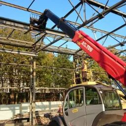 Montaż stalowej konstrukcji hali przez ekipę z wykorzystaniem podnośnika teleskopowego Manitou. Widoczne drzewa w tle.