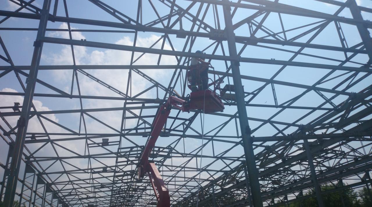 Pracownik na podnośniku koszowym montuje stalową konstrukcję dachu na tle błękitnego nieba z chmurami. Widok z dołu ukazuje skomplikowaną siatkę belek.