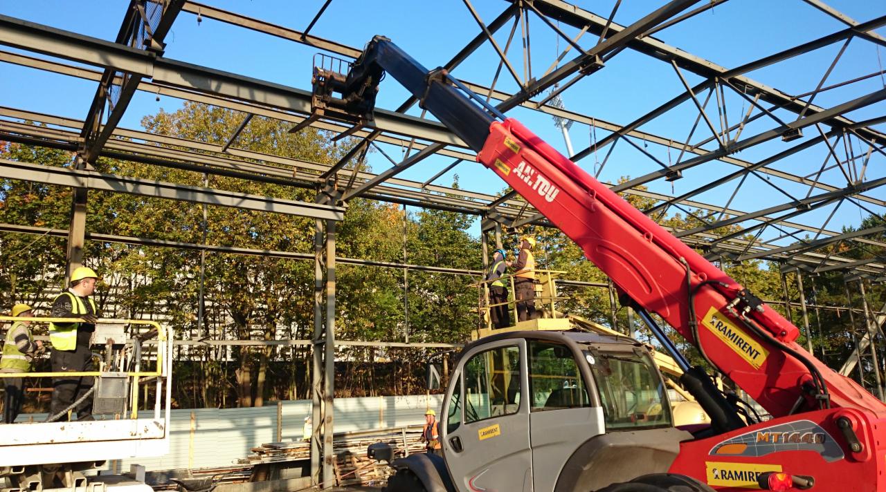 Montaż stalowej konstrukcji hali przez ekipę z wykorzystaniem podnośnika teleskopowego Manitou. Widoczne drzewa w tle.